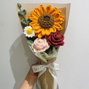 Könnte beinhalten: Ein gehäkelter Blumenstrauß in einer Juteschachtel mit einem weißen Band, auf dem "Liebe ist ewig" steht. Der Strauß enthält eine große gelbe Sonnenblume, eine rosa Rose, eine rote Rose und eine weiße Gänseblümchen.