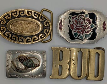May include: Four belt buckles: an oval brass buckle with a Greek key border, a rectangular silver-tone buckle with a red and green rose inlay, a small rectangular silver-tone buckle with a stone accent, and a large brass buckle spelling BUD.