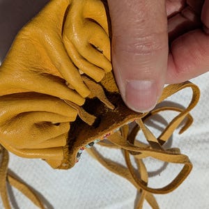 Puede incluir: Bolsa de cuero amarillo mostaza con cierre de cord&oacute;n. La bolsa est&aacute; abierta, revelando un borde con cuentas. El cord&oacute;n est&aacute; hecho de finas tiras de cuero marr&oacute;n. La bolsa est&aacute; hecha de cuero suave y texturizado.