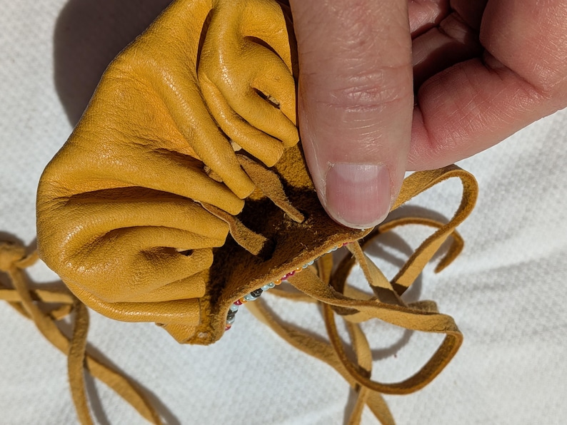 Puede incluir: Bolsa de cuero amarillo mostaza con cierre de cord&oacute;n. La bolsa est&aacute; abierta, revelando un borde con cuentas. El cord&oacute;n est&aacute; hecho de finas tiras de cuero marr&oacute;n. La bolsa est&aacute; hecha de cuero suave y texturizado.