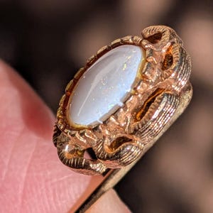 Op de afbeelding: Een vintage goudkleurige reversspeld met een ovale, iriserende witte steen. De steen is gezet in een gedetailleerde, sierlijke lijst. De speld wordt onder een hoek gehouden, waarbij het ingewikkelde ontwerp en het kleurenspel van de steen te zien zijn.