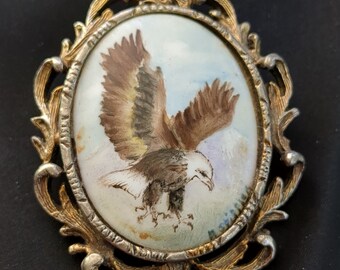 May include: A gold-toned metal frame with an oval-shaped glass insert. The glass insert features a hand-painted image of a brown and white eagle in flight.