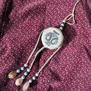 Puede incluir: Un colgante crema con un diseño tribal negro, cuentas blancas y negras y tres conchas de cauri. El colgante cuelga de un cordón crema sobre un fondo floral burdeos.