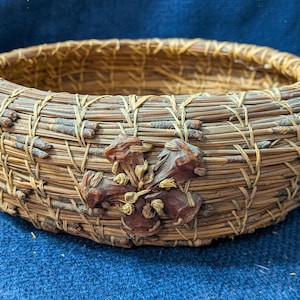 Puede incluir: Una cesta redonda construida con agujas de pino marrones enrolladas y ramitas finas, con un acento decorativo en forma de flor hecho de piezas de madera marr&oacute;n oscuro unidas a la pared exterior con costuras de fibra natural clara.