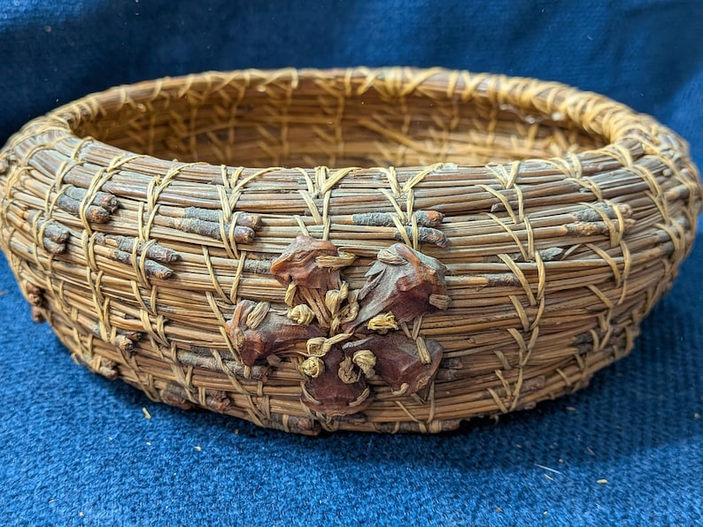 Puede incluir: Una cesta redonda construida con agujas de pino marrones enrolladas y ramitas finas, con un acento decorativo en forma de flor hecho de piezas de madera marr&oacute;n oscuro unidas a la pared exterior con costuras de fibra natural clara.