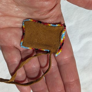 Puede incluir: Peque&ntilde;a bolsa cuadrada de cuero marr&oacute;n con un borde de cuentas rojas, amarillas, azules y negras. Una correa de cuero marr&oacute;n se extiende desde la parte inferior. La bolsa se sostiene en una mano.