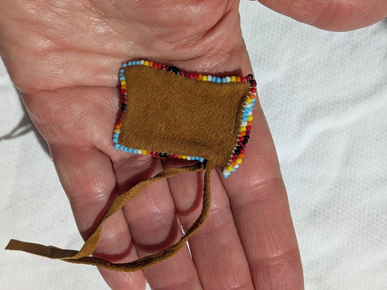 Puede incluir: Peque&ntilde;a bolsa cuadrada de cuero marr&oacute;n con un borde de cuentas rojas, amarillas, azules y negras. Una correa de cuero marr&oacute;n se extiende desde la parte inferior. La bolsa se sostiene en una mano.