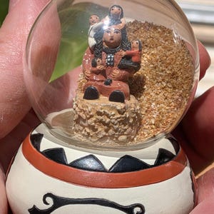 May include: A snow globe featuring a Native American woman with three children. The figurine is atop a textured base, surrounded by sand, and encased in a clear glass globe. The base is white with black and orange geometric designs.