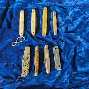 以下が含まれることがあります： さまざまなハンドルデザインとブレード形状の小さなポケットナイフのコレクション。白い、黄色、または茶色のハンドルが付いたナイフもあります。装飾的な銀色のハンドルが付いたナイフが1つあります。ナイフは青いベルベットの表面に配置されています。