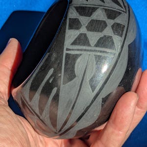 Pode incluir: Uma tigela de cerâmica preta polida com padrões geométricos e folhagens gravados. A tigela tem formato arredondado e uma abertura larga. Os desenhos gravados são em um tom mais claro, criando um contraste com a superfície escura.