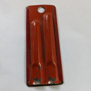 May include: A red metal object with two slots and a hole at the top. The object is slightly rusted and has a worn appearance.