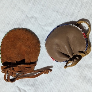 Puede incluir: Dos peque&ntilde;as bolsas de cuero marr&oacute;n con bordes de cuentas y cierres de cord&oacute;n. Una bolsa es de color marr&oacute;n oscuro con cuentas verdes, la otra es de color marr&oacute;n claro con cuentas rojas, blancas y azules. Ambas tienen un lazo.