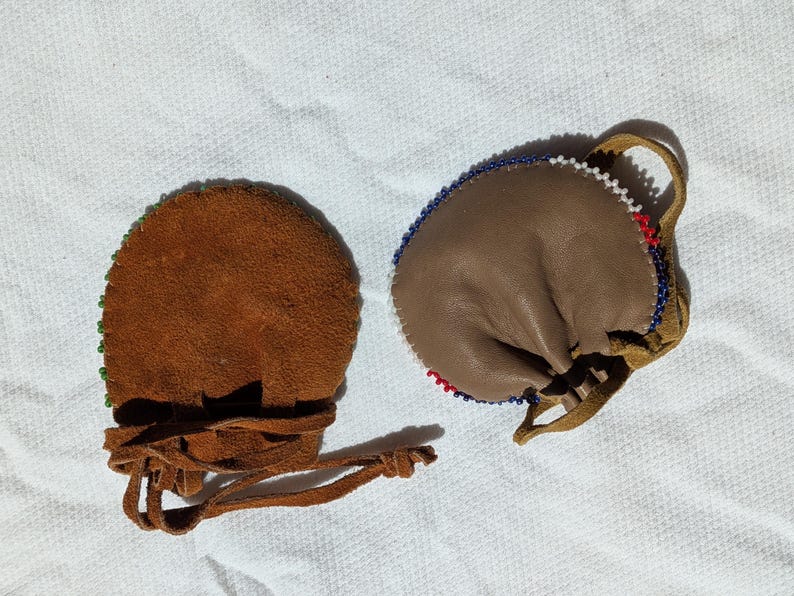 Puede incluir: Dos peque&ntilde;as bolsas de cuero marr&oacute;n con bordes de cuentas y cierres de cord&oacute;n. Una bolsa es de color marr&oacute;n oscuro con cuentas verdes, la otra es de color marr&oacute;n claro con cuentas rojas, blancas y azules. Ambas tienen un lazo.