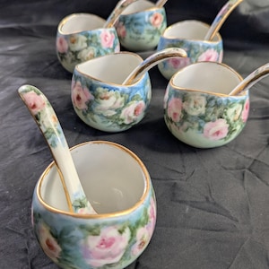 Peut inclure: Un ensemble de six petits bols en porcelaine avec cuillères assorties, ornés d'un motif floral peint à la main de roses roses et de feuilles vertes sur fond bleu clair, avec un fin liseré doré sur les bords des bols et des cuillères.