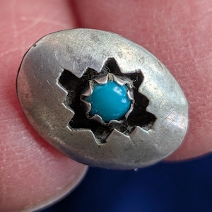 Puede incluir: Pendiente de botón ovalado plateado con una piedra turquesa en el centro. El pendiente tiene un diseño de estrella alrededor de la piedra turquesa. El pendiente se sostiene entre dos dedos.