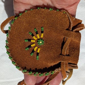 Puede incluir: Una bolsa de gamuza marr&oacute;n con un dise&ntilde;o floral de cuentas. La bolsa est&aacute; decorada con cuentas verdes, amarillas y negras alrededor de los bordes y en la parte delantera. Tiene un lazo y flecos.
