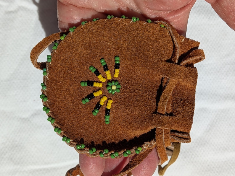 Puede incluir: Una bolsa de gamuza marr&oacute;n con un dise&ntilde;o floral de cuentas. La bolsa est&aacute; decorada con cuentas verdes, amarillas y negras alrededor de los bordes y en la parte delantera. Tiene un lazo y flecos.