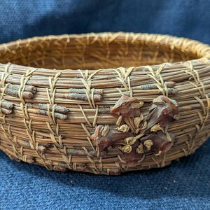 Puede incluir: Una cesta redonda y poco profunda hecha de agujas de pino enrolladas en tonos marr&oacute;n y beige. El exterior presenta un segmento decorativo de pi&ntilde;a de color marr&oacute;n oscuro fijado en el lateral, con peque&ntilde;as ramitas tejidas en las agujas.