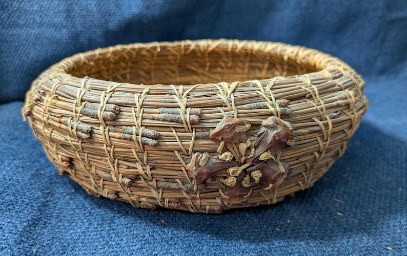 Puede incluir: Una cesta redonda y poco profunda hecha de agujas de pino enrolladas en tonos marr&oacute;n y beige. El exterior presenta un segmento decorativo de pi&ntilde;a de color marr&oacute;n oscuro fijado en el lateral, con peque&ntilde;as ramitas tejidas en las agujas.
