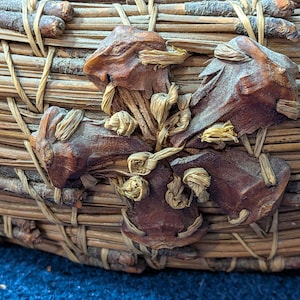 Puede incluir: Una cesta circular construida con agujas de pino marrones tejidas y peque&ntilde;as ramitas. El exterior presenta un elemento decorativo prominente hecho de piezas de madera de color marr&oacute;n oscuro en forma de estrella fijadas con fibras de rafia.