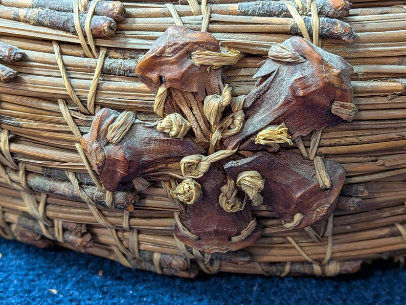 Puede incluir: Una cesta circular construida con agujas de pino marrones tejidas y peque&ntilde;as ramitas. El exterior presenta un elemento decorativo prominente hecho de piezas de madera de color marr&oacute;n oscuro en forma de estrella fijadas con fibras de rafia.