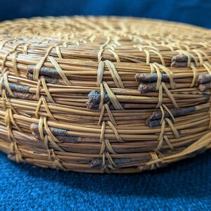 Puede incluir: Una cesta redonda y plana construida con agujas de pino enrolladas y peque&ntilde;as ramitas de color marr&oacute;n oscuro. Las agujas est&aacute;n unidas con un hilo de fibra natural claro en un patr&oacute;n de puntada repetitivo, creando una superficie circular texturizada.