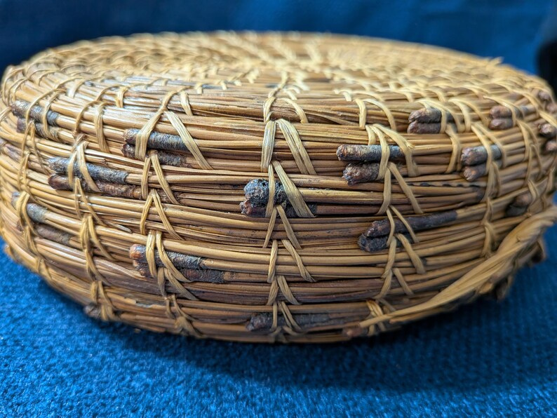 Puede incluir: Una cesta redonda y plana construida con agujas de pino enrolladas y peque&ntilde;as ramitas de color marr&oacute;n oscuro. Las agujas est&aacute;n unidas con un hilo de fibra natural claro en un patr&oacute;n de puntada repetitivo, creando una superficie circular texturizada.