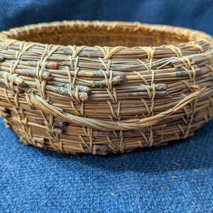 Puede incluir: Una cesta redonda y poco profunda construida con agujas de pino enrolladas y peque&ntilde;as ramitas finas. Las agujas de color marr&oacute;n y bronceado natural est&aacute;n unidas con hilo claro en un patr&oacute;n de puntada visible y repetitivo, creando un recipiente circular.
