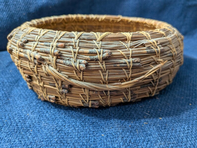 Puede incluir: Una cesta redonda y poco profunda construida con agujas de pino enrolladas y peque&ntilde;as ramitas finas. Las agujas de color marr&oacute;n y bronceado natural est&aacute;n unidas con hilo claro en un patr&oacute;n de puntada visible y repetitivo, creando un recipiente circular.