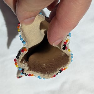 Puede incluir: Peque&ntilde;a bolsa abierta de cuero color canela, bordeada con cuentas de colores. Las cuentas forman un patr&oacute;n de rojo, naranja, amarillo, negro, blanco y azul. La bolsa se sostiene abierta, revelando el interior.