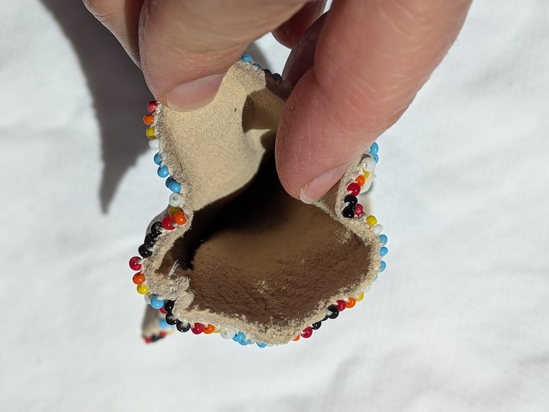 Puede incluir: Peque&ntilde;a bolsa abierta de cuero color canela, bordeada con cuentas de colores. Las cuentas forman un patr&oacute;n de rojo, naranja, amarillo, negro, blanco y azul. La bolsa se sostiene abierta, revelando el interior.