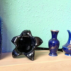 Könnte beinhalten: Sammlung blauer dekorativer Keramikartikel. Im Vordergrund ein sternförmiger schwarzer Aschenbecher, dazu eine kleine blaue Vase mit goldenem Rand und Text sowie ein blauer Krug mit goldenen floralen Details.