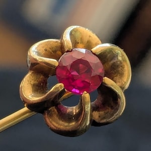May include: A gold-toned floral pin with a vibrant, faceted ruby gemstone at its center. The pin's petals are detailed and curved, creating a three-dimensional effect. The ruby is a deep, rich red, and the pin has a classic, vintage aesthetic.
