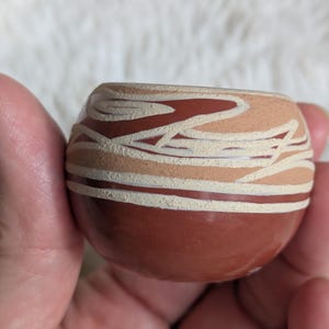 May include: A small, round, hand-painted ceramic pot. The pot features a dark reddish-brown base with a lighter tan and white design around the upper portion. The design includes curved lines and shapes, creating a unique pattern.