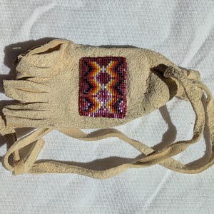 Puede incluir: Una bolsa de cuero beige con un dise&ntilde;o de cuentas en tonos rojos, naranjas, amarillos y morados. La bolsa tiene un borde con flecos y un cierre de cord&oacute;n. El dise&ntilde;o de cuentas es un patr&oacute;n geom&eacute;trico.