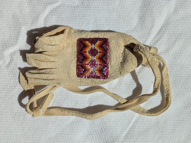 Puede incluir: Una bolsa de cuero beige con un dise&ntilde;o de cuentas en tonos rojos, naranjas, amarillos y morados. La bolsa tiene un borde con flecos y un cierre de cord&oacute;n. El dise&ntilde;o de cuentas es un patr&oacute;n geom&eacute;trico.