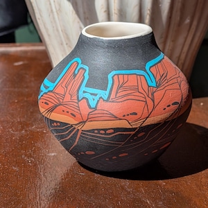May include: A small, round ceramic vase with a black exterior and a white interior. The vase features a landscape design in shades of red, orange, and blue. The design depicts a desert scene with mountains and mesas.