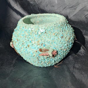 Puede incluir: Una pequeña maceta decorativa redonda de color turquesa. El exterior está cubierto de pequeños trozos texturizados, lo que le da un aspecto rugoso. La maceta tiene una amplia abertura y está acentuada con detalles marrones y rosas.