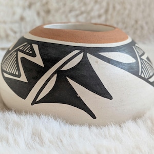 Può includere: Una ciotola in ceramica bianco sporco fatta a mano, con un'ampia apertura e un bordo marrone. La ciotola è adornata da audaci motivi geometrici neri, tra cui triangoli e linee. La ciotola è appoggiata su una superficie morbida e bianca.