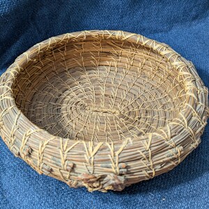 Puede incluir: Una cesta redonda y poco profunda hecha de agujas de pino enrolladas. Las agujas de color marr&oacute;n natural est&aacute;n unidas con fibras finas de color claro en un patr&oacute;n de puntadas repetitivo, creando una base circular texturizada y un borde elevado.