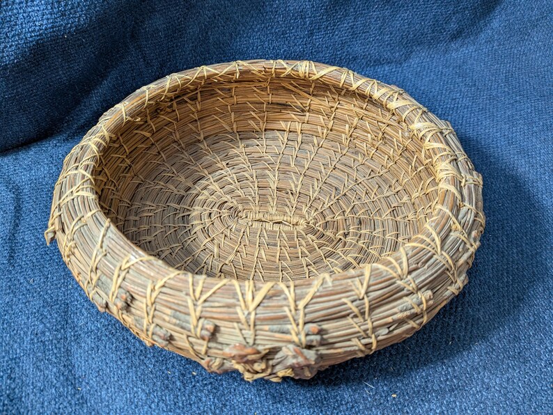 Puede incluir: Una cesta redonda y poco profunda hecha de agujas de pino enrolladas. Las agujas de color marr&oacute;n natural est&aacute;n unidas con fibras finas de color claro en un patr&oacute;n de puntadas repetitivo, creando una base circular texturizada y un borde elevado.