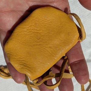 Puede incluir: Una peque&ntilde;a bolsa de cuero amarillo mostaza con cierre de cord&oacute;n. La bolsa es rectangular con bordes redondeados y una superficie texturizada. Se adjunta un fino cord&oacute;n de cuero para llevar.