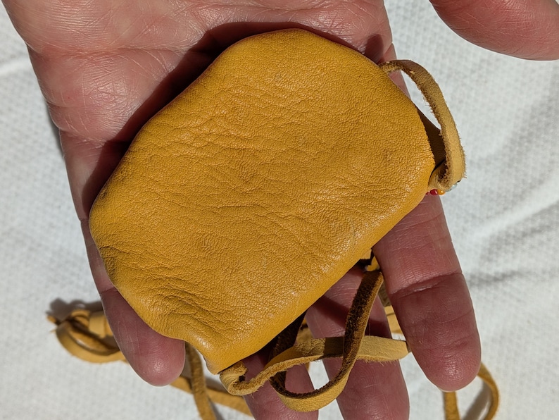 Puede incluir: Una peque&ntilde;a bolsa de cuero amarillo mostaza con cierre de cord&oacute;n. La bolsa es rectangular con bordes redondeados y una superficie texturizada. Se adjunta un fino cord&oacute;n de cuero para llevar.