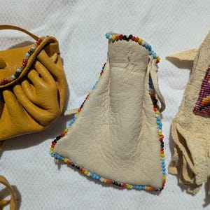 Puede incluir: Primer plano de tres bolsas de cuero hechas a mano. Una es de color amarillo mostaza, otra es de color blanco roto con ribete de cuentas de colores, y la tercera es de color beige claro con un dise&ntilde;o de cuentas. Las bolsas son probablemente para llevar objetos peque&ntilde;os.