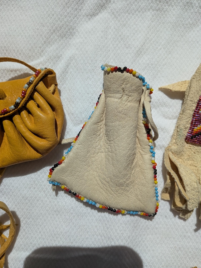 Puede incluir: Primer plano de tres bolsas de cuero hechas a mano. Una es de color amarillo mostaza, otra es de color blanco roto con ribete de cuentas de colores, y la tercera es de color beige claro con un dise&ntilde;o de cuentas. Las bolsas son probablemente para llevar objetos peque&ntilde;os.