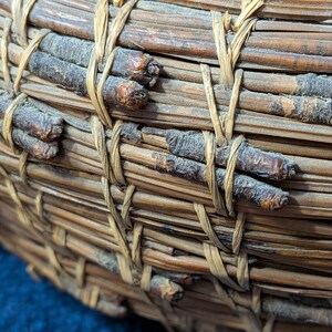 Puede incluir: Una cesta tejida a mano hecha de agujas de pino marrones y ramitas oscuras y delgadas. La estructura presenta capas horizontales de agujas de pino agrupadas, aseguradas por ataduras verticales de material vegetal fibroso color canela.