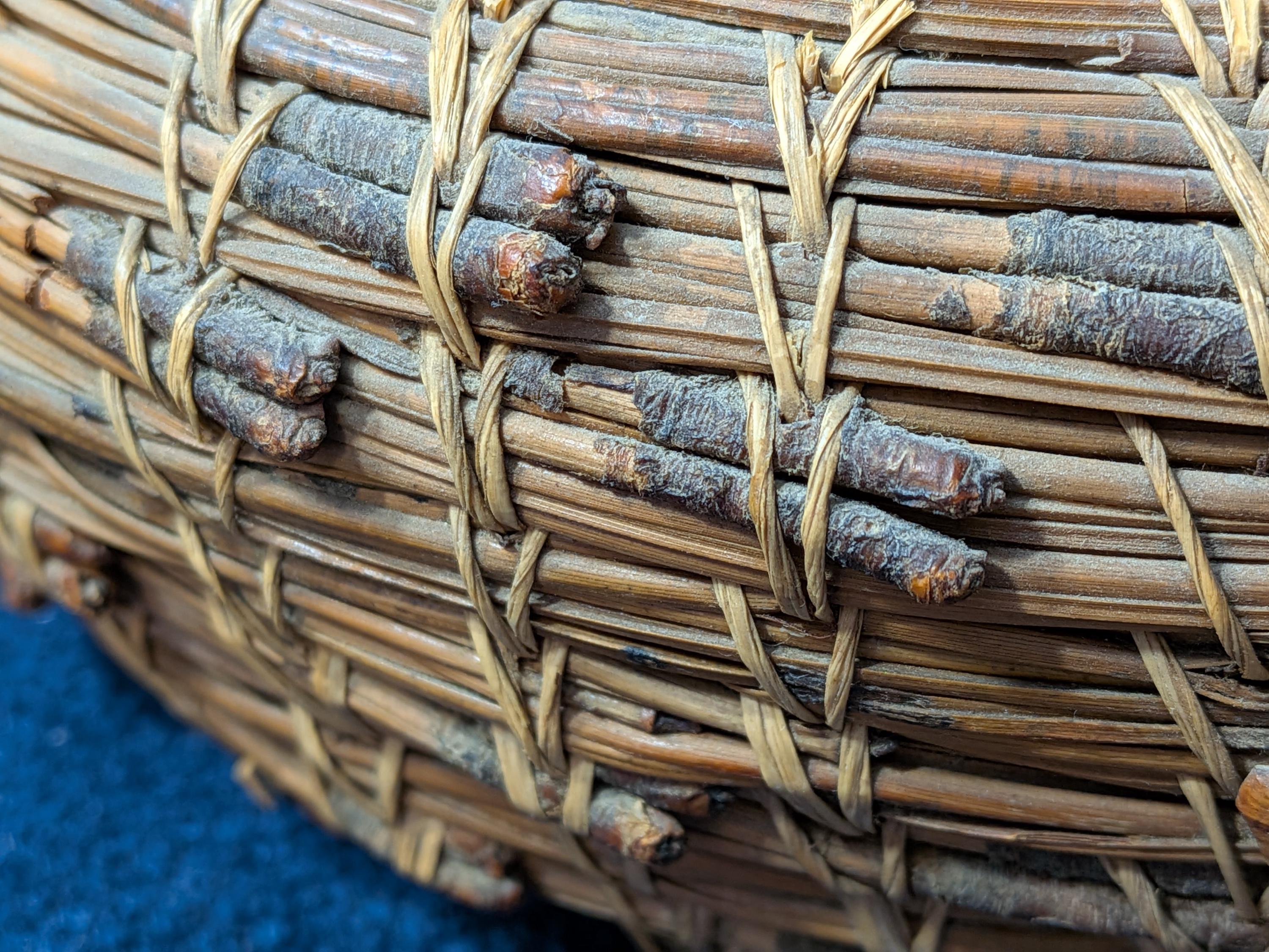 Puede incluir: Una cesta tejida a mano hecha de agujas de pino marrones y ramitas oscuras y delgadas. La estructura presenta capas horizontales de agujas de pino agrupadas, aseguradas por ataduras verticales de material vegetal fibroso color canela.