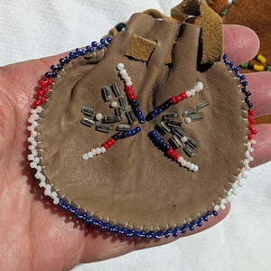 Puede incluir: Una peque&ntilde;a bolsa redonda de cuero marr&oacute;n con cierre de cord&oacute;n. La bolsa est&aacute; decorada con un dise&ntilde;o de cuentas en rojo, blanco y azul. El borde de la bolsa tambi&eacute;n est&aacute; adornado con cuentas blancas, azules y rojas.