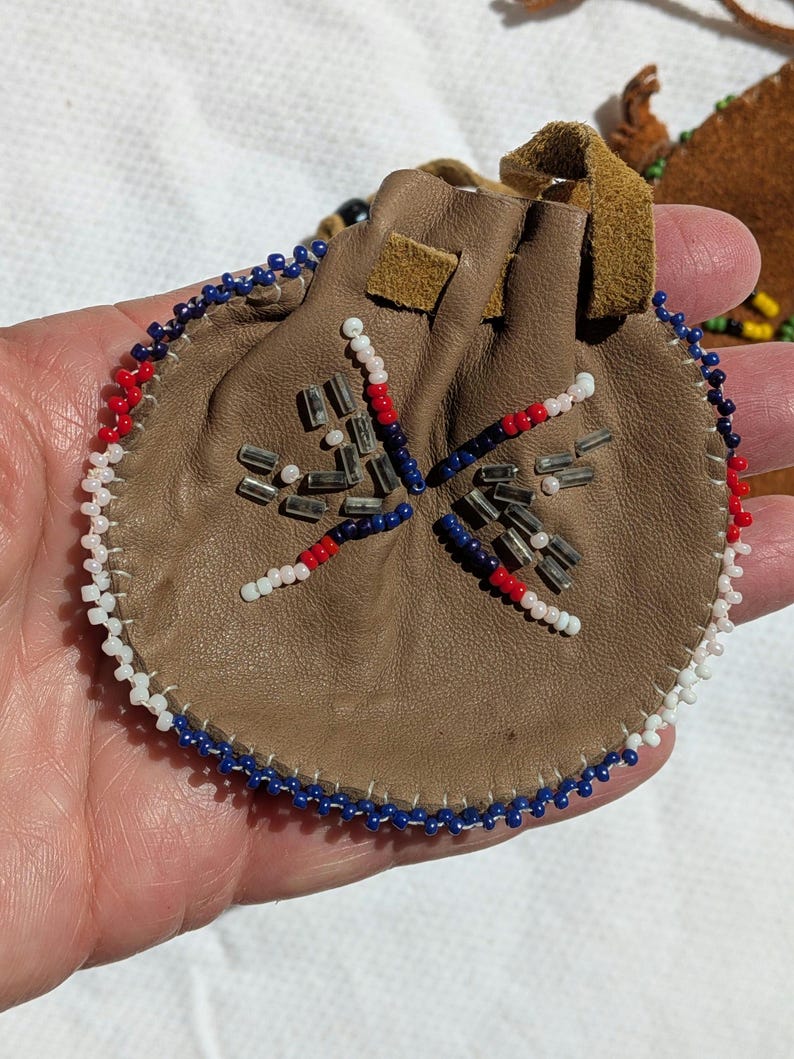 Puede incluir: Una peque&ntilde;a bolsa redonda de cuero marr&oacute;n con cierre de cord&oacute;n. La bolsa est&aacute; decorada con un dise&ntilde;o de cuentas en rojo, blanco y azul. El borde de la bolsa tambi&eacute;n est&aacute; adornado con cuentas blancas, azules y rojas.