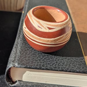 Peut inclure: Un petit bol rond en poterie de couleur terre cuite avec une large ouverture. Le bol est décoré de bandes horizontales de couleurs rouge foncé et beige alternées, avec un motif géométrique. Le bol repose sur un livre relié en cuir noir.